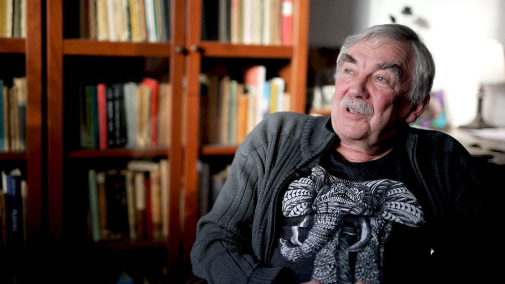 Portret mężczyzny w koszuli ze słoniemi i swetrze, w tle półka z ksiażkami