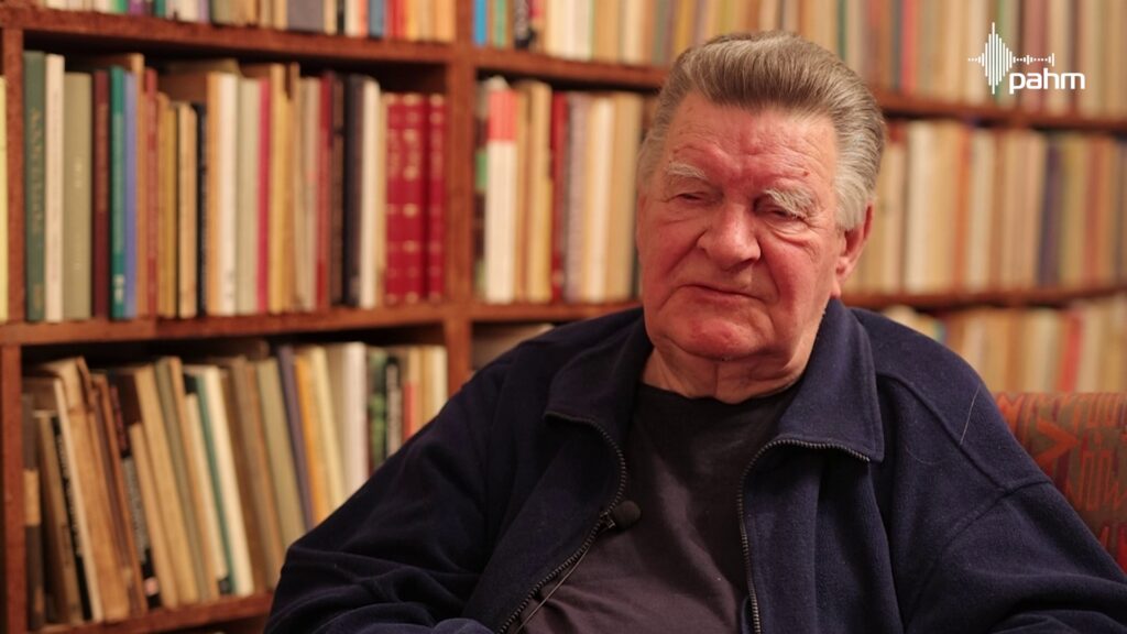 Mężczyzna w ciemnej bluzie siedzi i opowiada. Za nim w tle półka z książkami.